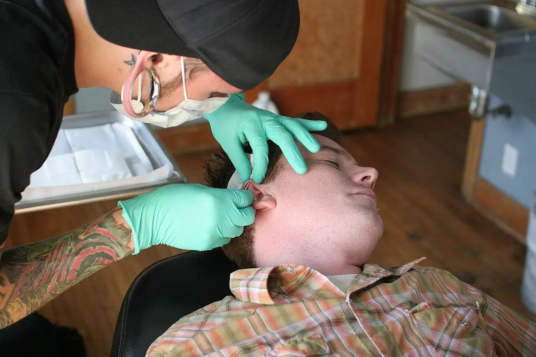 A masked piercer in nitrile gloves holding a client's ear to pierce the helix, with a covered tray beside the chair