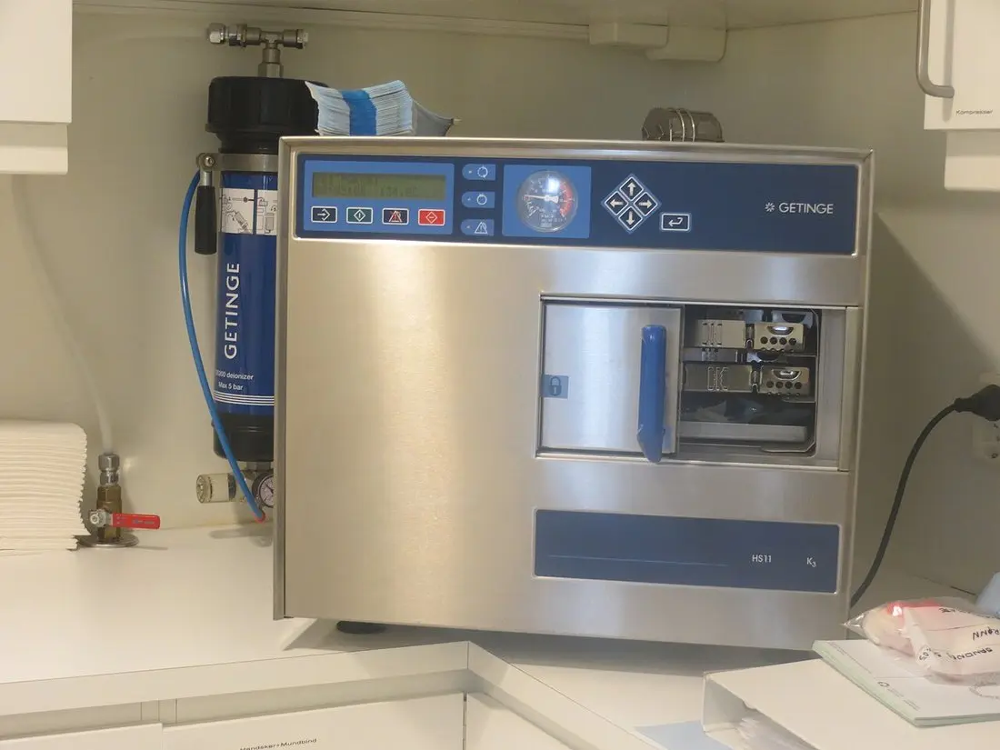 A stainless-steel tabletop steam autoclave with its door open and instrument trays inside, in a clinic